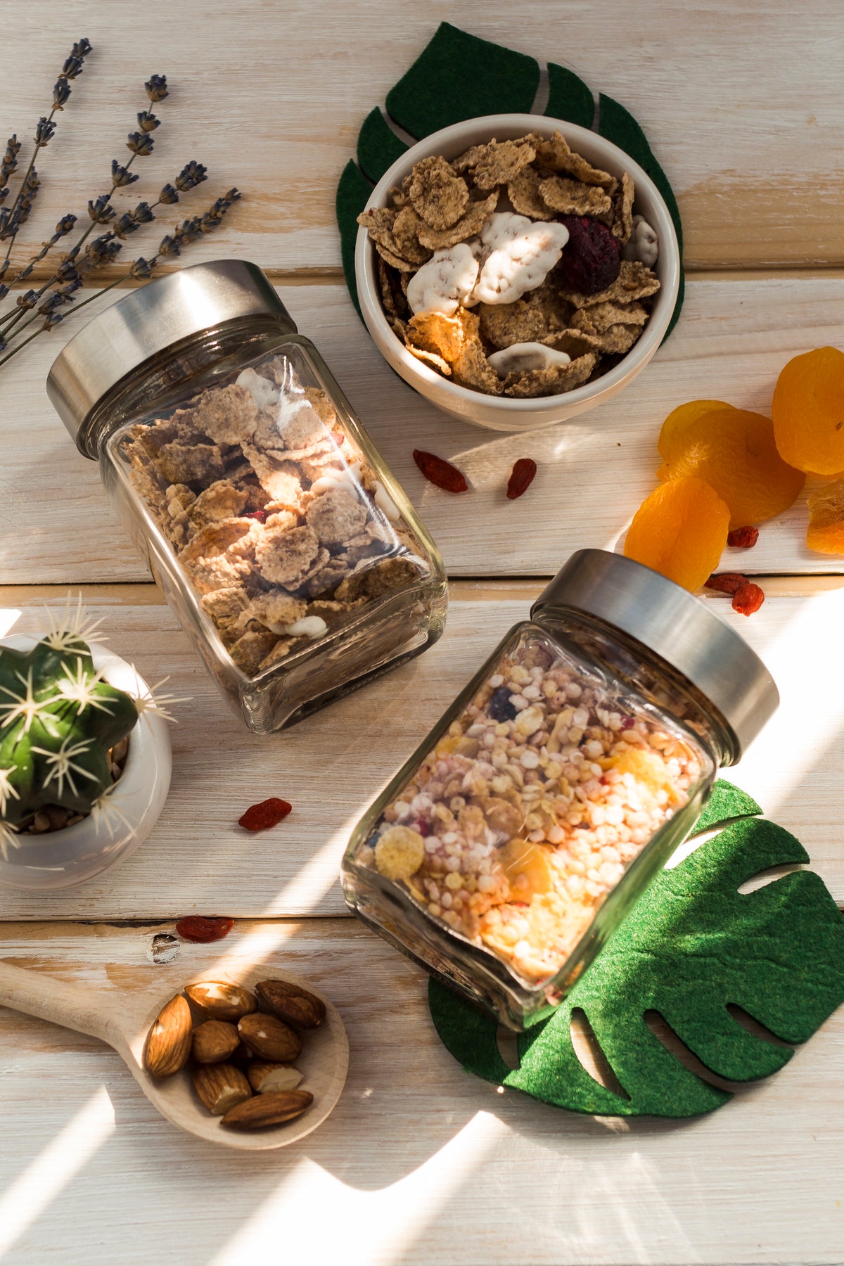 Two glass jars filled with various seeds and grains on a wooden surface with a bowl of cereal and dried fruits.